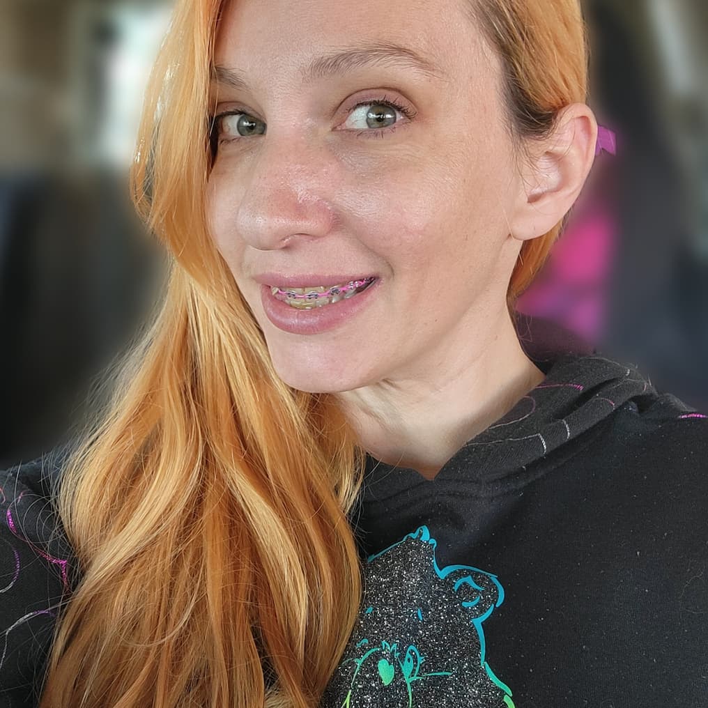 [SYSTEM_LOG: FULL_COLOR_RECOVERY] Close-up headshot of Kellye Strickland, a woman with long strawberry-blonde hair and green eyes. She is smiling, showing orthodontic braces with pink bands. She is wearing a black hoodie with a glittery Care Bear graphic. The background is softly blurred.